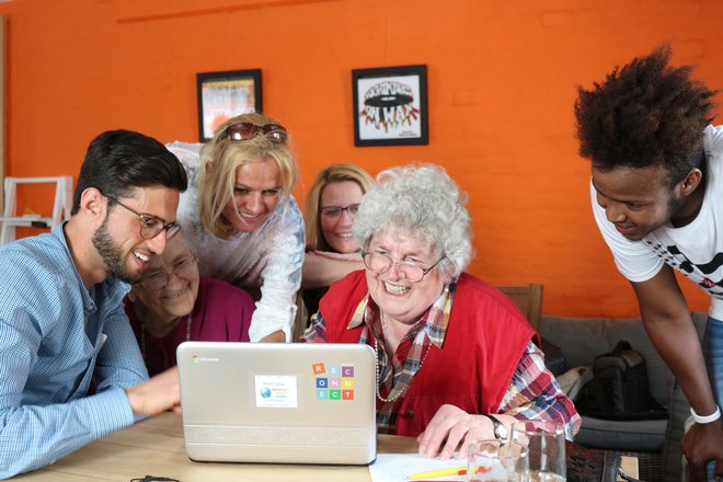Junge Menschen sitzen und stehen um eine Seniorin herum und helfen ihr bei der Eingabe im vor ihr stehenden Laptop.