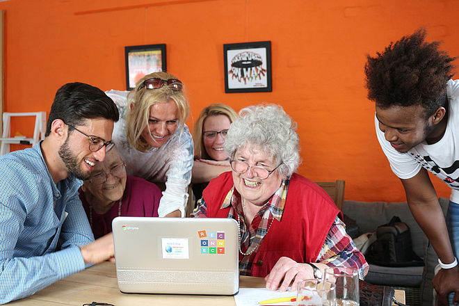 Junge Menschen sitzen und stehen um eine Seniorin herum und helfen ihr bei der Eingabe im vor ihr stehenden Laptop.