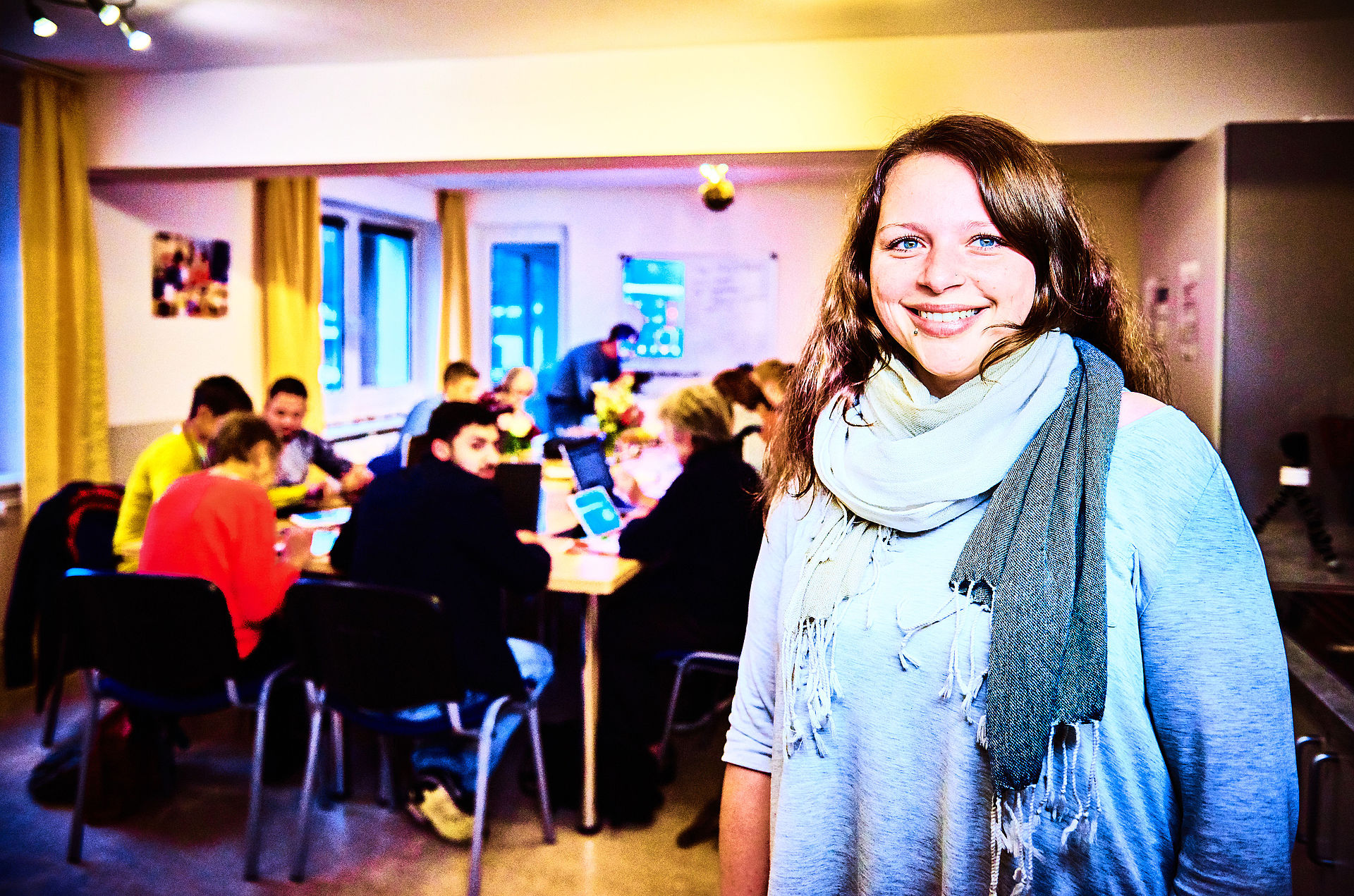 eine junge Frau steht im Vordergrund und lächelt in die Kamera, im Hintergrund sitzen Menschen an einem Tisch