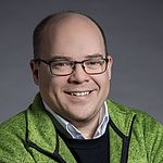 Porträt von einem Mann, der eine grüne Jacke trägt und in die Kamera lächelt.