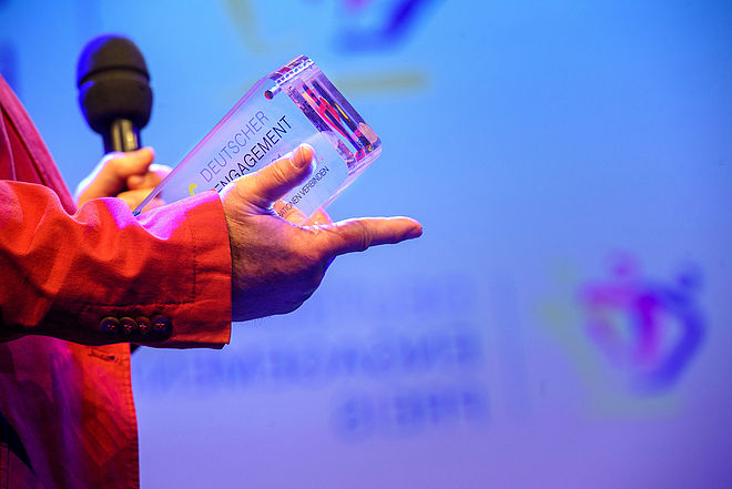 Eine Hand hält einen Pokal seitlich ins Bild, das Foto wurde auf einer Bühne aufgenommen und ist in Blautönen gehalten