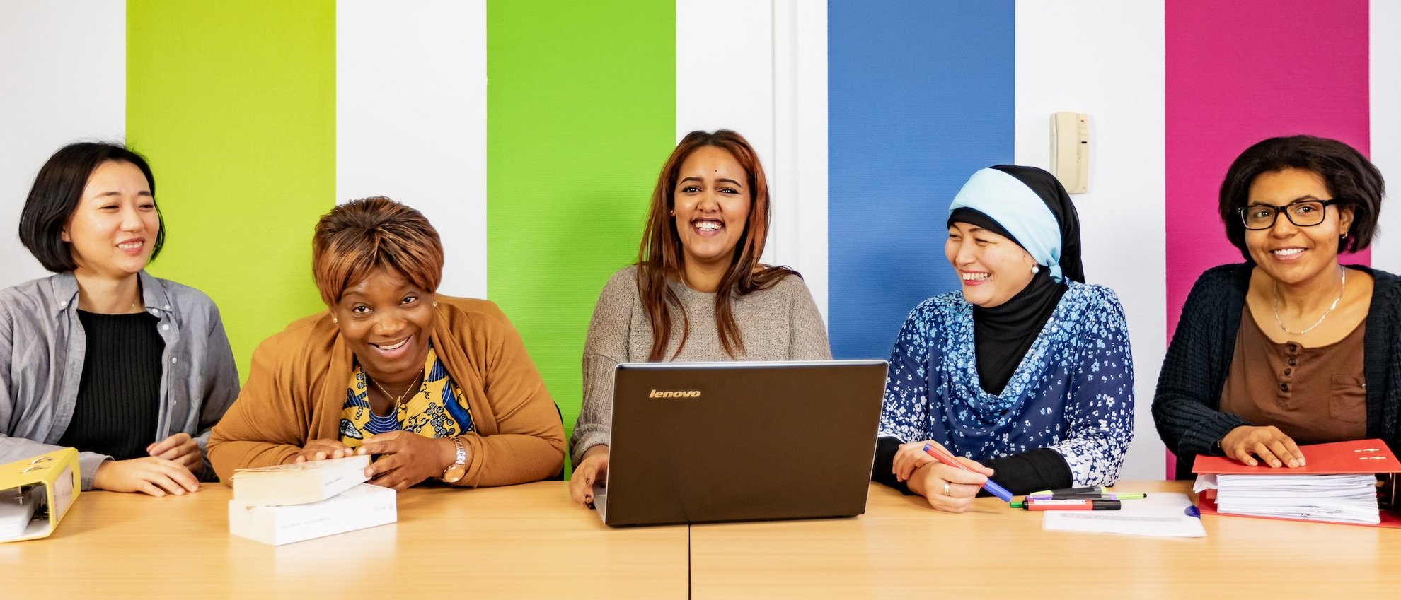 Fünf Frauen sitzen vor einer bunt gestreiften Wand an einem Tisch mit Laptops und Ordnern