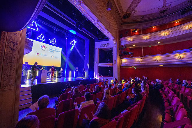 Der Theatersaal des Deutschen Theaters Berlin, das Publikum schaut auf die Bühne, wo man Moderatorin Jana Pareigis vor der Leinwand des Deutschen Engagementpreises sieht.