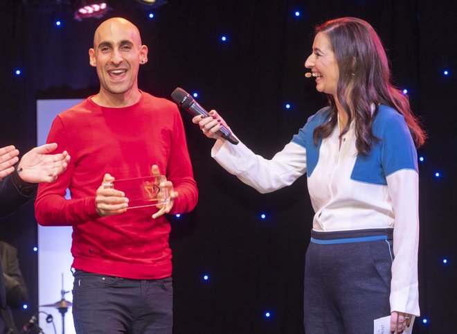 Ein Preisträger (mittig) steht auf der Bühne mit dem Pokal des Deutschen Engagementpreises, rechts davon die Moderatorin, die ihn interviewt und auf der linken Seite ein applaudierender Mann.