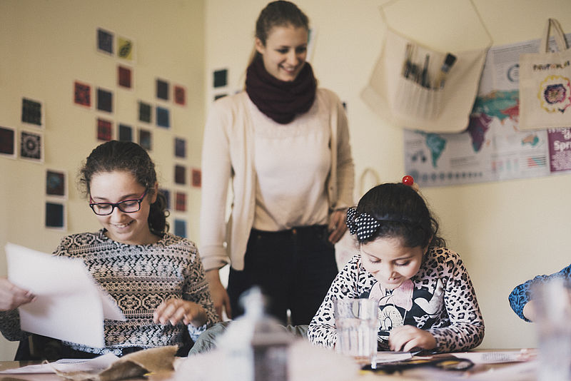 Zwei Kinder arbeiten zusammen mit einer Ehrenamtlichen an Kunst-Projekten