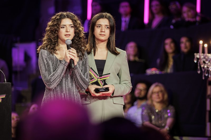 Jihan hält ein Mikrofon, um sie herum Publikum, neben ihr ihre Schwester, die weinend ihren Award hält