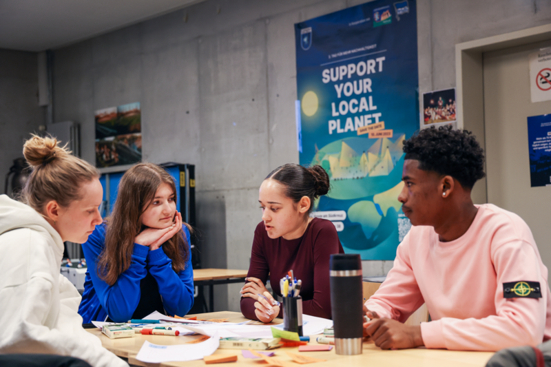 Eine Person steht in der Mitte des Bilds und präsentiert, während insgesamt sieben sichtbare Personen um ihn herum in einem Raum sind.