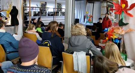 Mehrere Personen sitzen auf Stühlen und schauen zu einer Person, sie vorne steht und redet