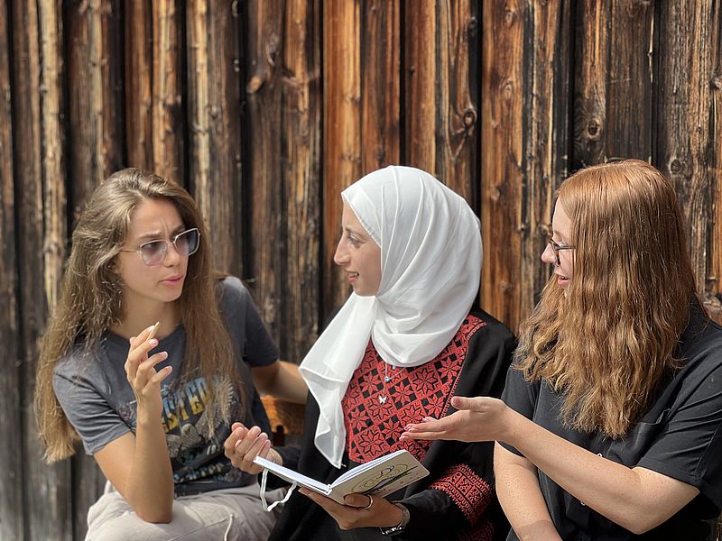 Auf dem Bild sieht man drei junge Frauen im angeregten Gespräch. In der Mitte sitzt eine Frau mit Kopftuch, die ein Buch in der Hand hält.