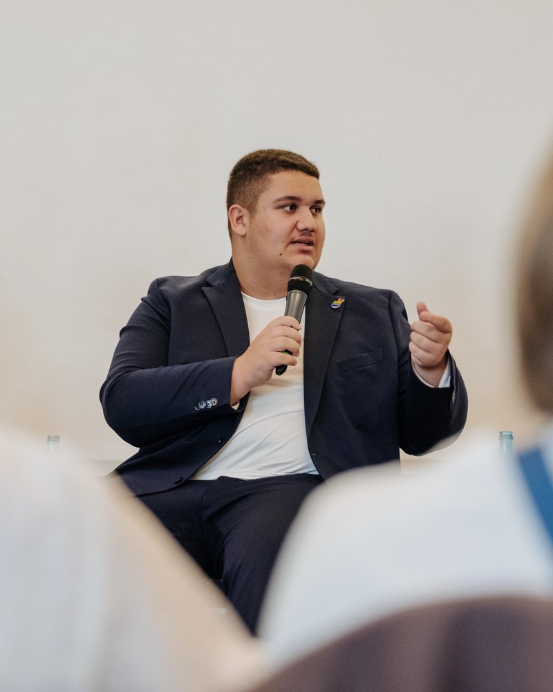 Max sitzt auf einem Podium und hat sein Kopf nach links gedreht. Er spricht. Das Mikrofon hält er in der rechten Hand, mit der linken gestikuliert er. Er trägt einen dunkelblauen Anzug und ein weißes T-Shirt darunter.