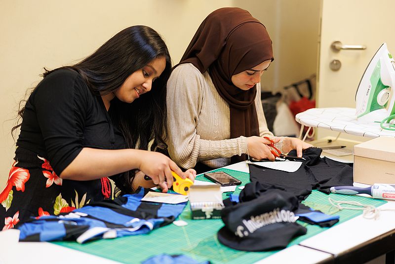 Zwei Frauen sitzen an einem Tisch voll mit Kleidungsutensilien, die repariert werden. Die linke Frau nutzt ein Etikettiergerät, die rechte Frau schneidet mit einer Schere einen Stoff zurecht.