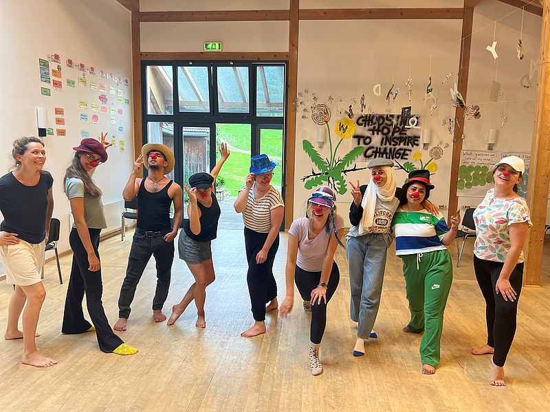 Auf dem Bild sieht man Teilnehmende der Sommerakademie mit roten Clownsnasen, die lustige Bewegungen zu machen scheinen Im Hintergrund sieht man das Motto der Sommerakademie: Chosing Hope to Inspire Change.