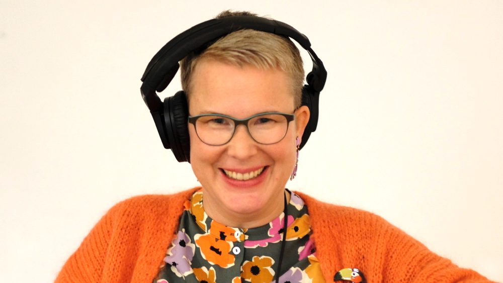 Eine Frau mit Kopfhörern und einer orangenen Jacke blickt freundlich in die Kamera und lächelt