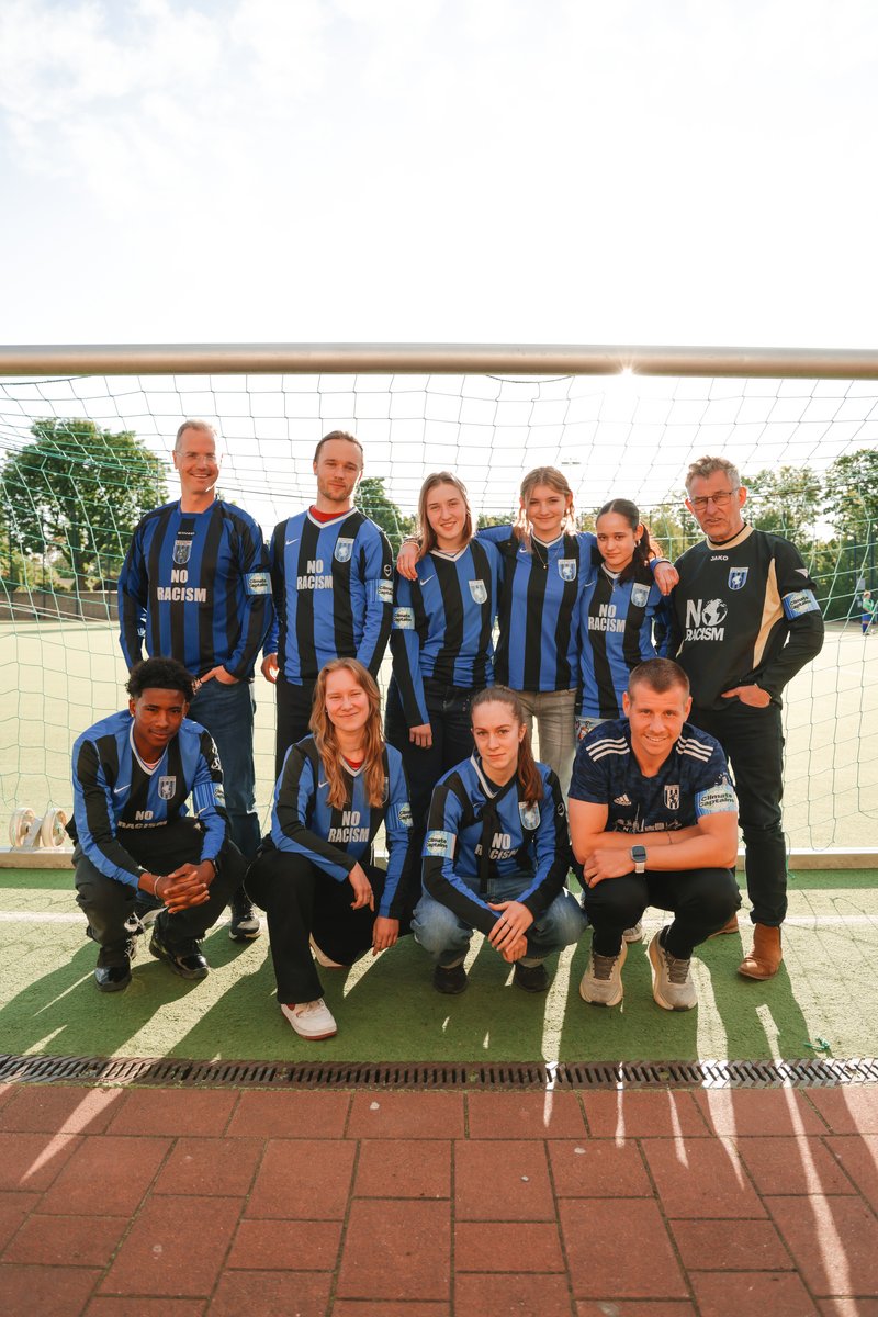6 stehende und vier hockende Menschen sind in zwei Reihen in einem Fußballtor positioniert und tragen blau schwarze Trikots
