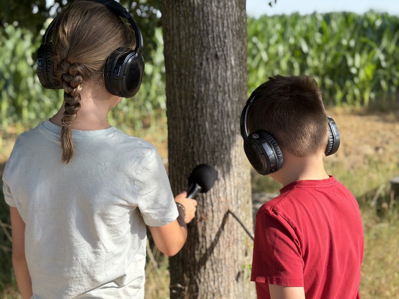 Zwei Kinder tragen Kopfhörer und nehmen mit einem Mikrofon die Geräusche eines Baumes auf.