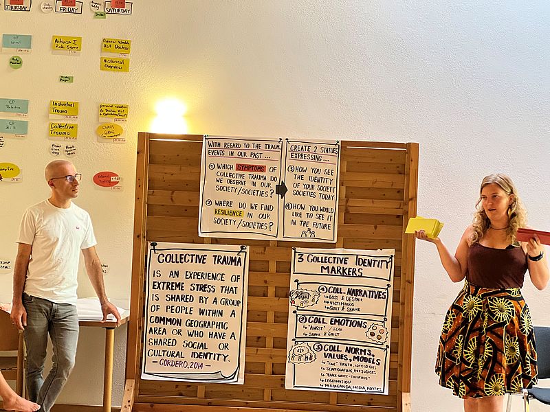 Auf dem Bild sieht man einen Referenten und eine Referentin links und rechts neben einer Pinnwand stehen. 