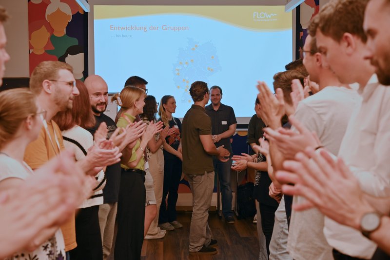 Junge Stotternde stehen vor einer PowerPoint Präsentation, mit dem Gesicht teils zur Präsentation / teils zur Kamera und klatschen. Die Mimik ist freudig. Auf der Präsentation sieht man die Entwicklung der Flow-Gruppe bis heute.