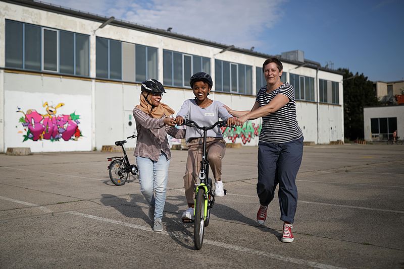 Eine Frau übt Fahrradfahren und wird von zwei anderen Frauen unterstützt 