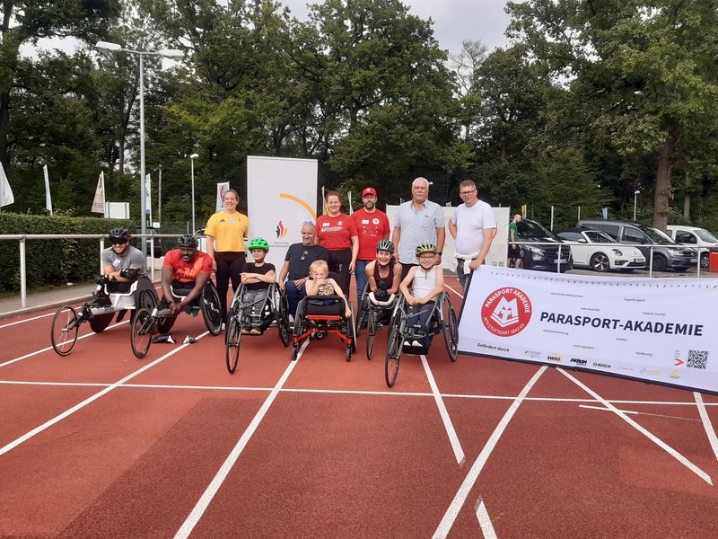 Sportler:innen im Rennrollstuhl stehen zum Foto auf der Tartanbahn. Hinter ihnen haben sich Trainer:innen und Funktionäre aufgestellt