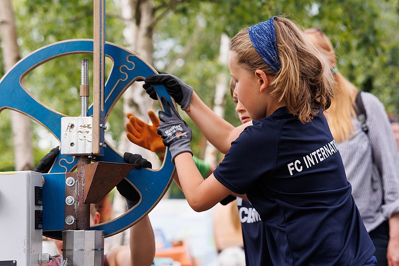 Ein Kind mit einem blauen FC Internationale Shirt wird von der Rückenansicht gezeigt, wie es eine Maschinen-Apparatur mit Händen in Arbeitshandschuhen dreht. Im Hintergrund schauen weitere Personen zu.