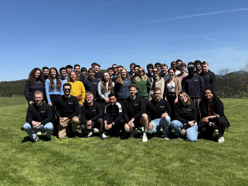 40 junge Menschen stehen und knien für ein Gruppenfoto auf einer Wiese vor blauem Himmel