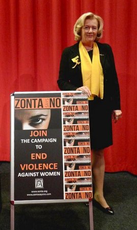 Renate von Köller mit gelbem Zonta-Schal steht neben einem Plakatständer der Kampagne „Zonta says NO" vor einem roten Vorhang