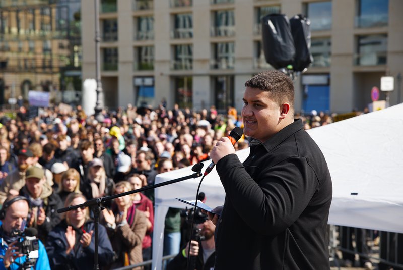 Max steht auf einer Bühne vor dem Brandenburger Tor. Er hält das Mikrofon in der linken Hand. Das Bild wurde von der Seite aufgenommen. Im Hintergrund sieht an viele Menschen, die klatschen.