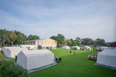 Mehrere Zelte und Bänke stehen auf einer Wiese auf einem Gelände. Menschen sitzen auf den Bänken oder stehen neben den Zelten.