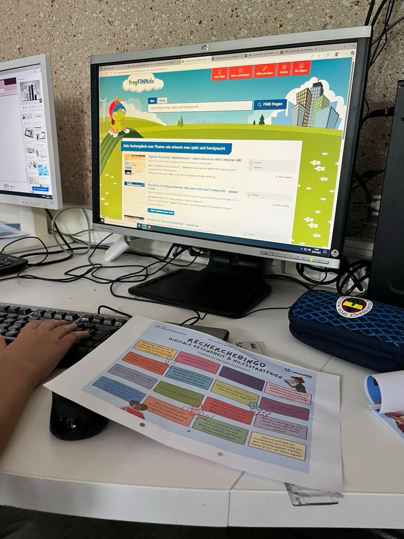 Ein Kind recherchiert am Computer mit der Kindersuchmaschine fragFINN. Auf dem Tisch liegt ein Recherche-Bingo zu digitalen Gesundheitsstrategien.