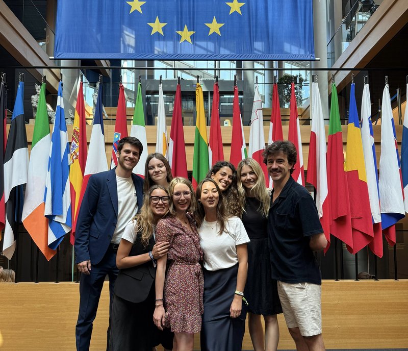 Acht junge Menschen stehen lächelnd vor Flaggen der EU
