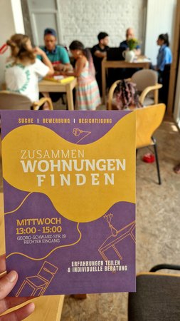 Im Vordergrund ein Infoflyer zum Workshop "Zusammen Wohnung finden", im Hintergrund sitzen Ehrenamtliche mit Wohnungssuchen an Tischen, Sie sind ins Gespräch vertieft, daneben spielen Kinder.