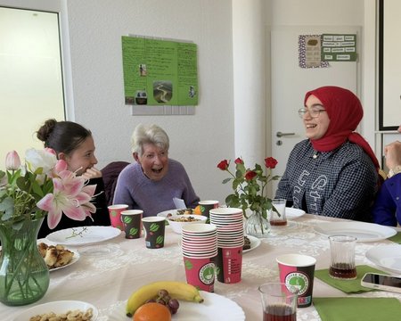 Seniorin und geflüchtete Jugendliche sitzen gemeinsam am Tisch und lachen miteinander.