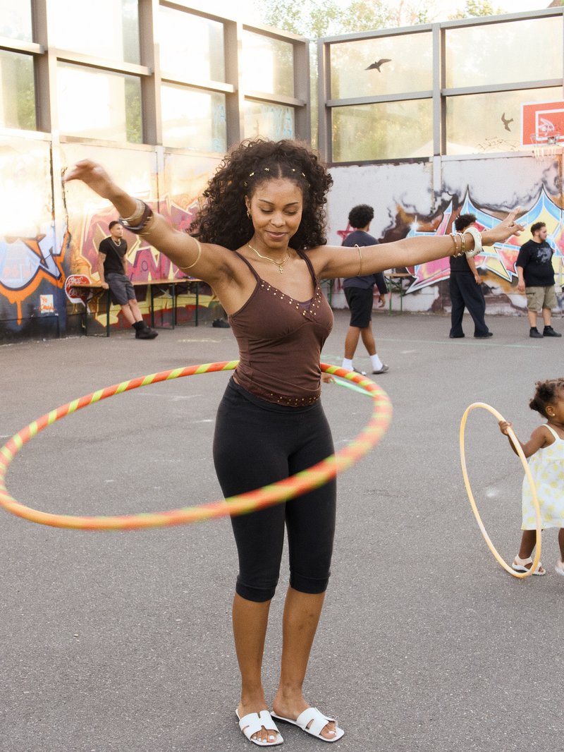 Eine Frau im Vordergrund die bei der Hula Hoop Workshop teilnimmt und lächelt, während im Hintergrund Jugendliche bei unsere Basketball Turnier während der Black Culture Garden spielen. 