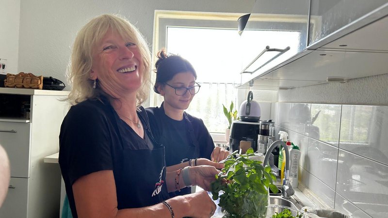 Eine deutsche Seniorin und eine geflüchtete Jugendliche lachen gemeinsam während eines Kochtreffens in einer Küche.