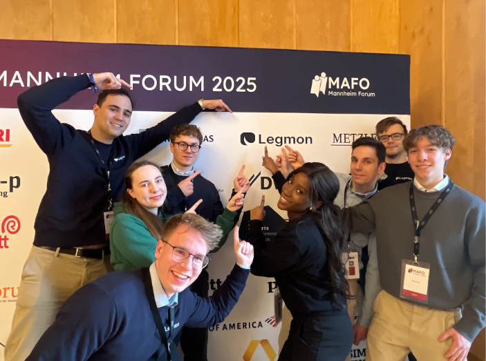 Acht Mitglieder des Legmon-Teams posieren lachend vor der Sponsorenwand des Mannheim Forum 2025