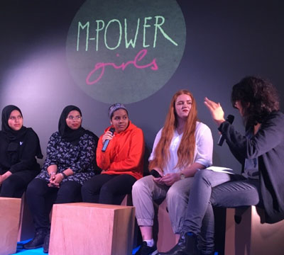 Eine Gruppe Mädchen sitzt vor einer Projektion des Mpower-Logos an einer Wand auf einer Bühne. Daneben sitzt eine Frau mit einem Mikrofon und gestikuliert mit den Händen. Auch eines der Mädchen hält ein Mikrofon. Alle blicken ernst aber freundlich zu der Frau. 