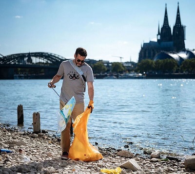  Ein Mann sammelt Müll am Rheinufer. Im Hintergrund der Kölner Dom und die Brücke.  
