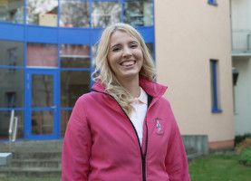 eine junge Frau in einer pinken Jacke mit Emblem der Kliniknannys lächelt