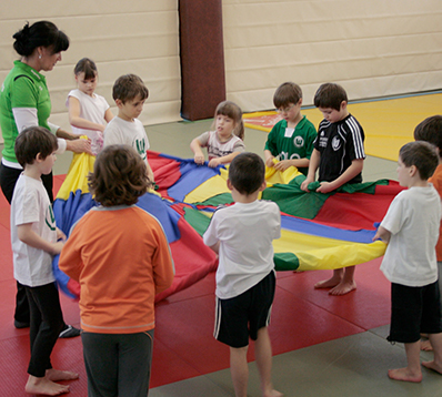  Erzieherin und Kinder im Kreis, halten buntes Tuch in einer Turnhalle